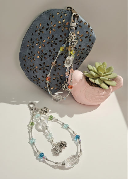 Two bag charms on a light surface with a decorative blue pouch and small pink pot with a succulent.